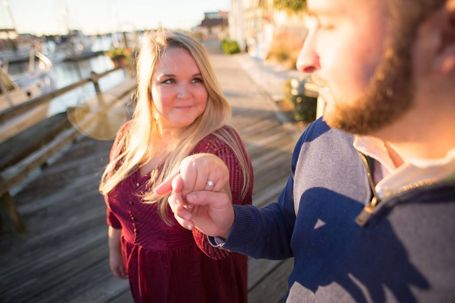 Woman smiles back at camera while holding partner's hand on a sunny waterfront boardwalk during engagement shoot.