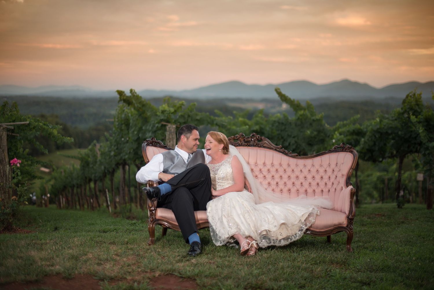 Bride and groom relaxing on an ornate pink velvet sofa in a vineyard during a golden sunset.