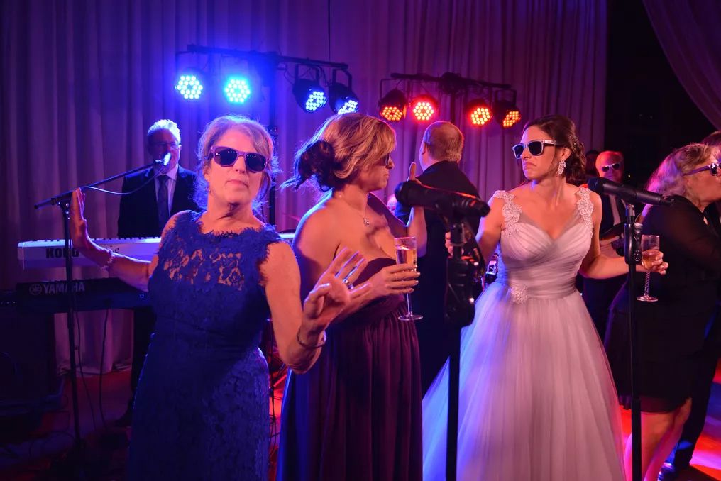 Bride in white gown dancing with guests wearing sunglasses at elegant wedding reception with stage lights.