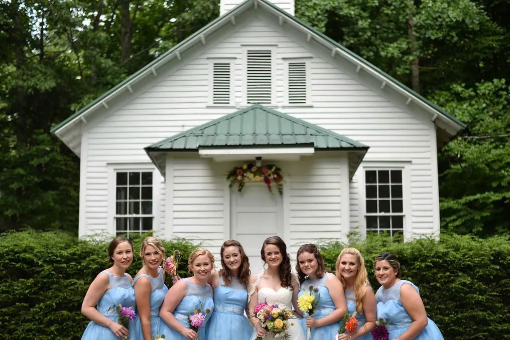Bride with seven bridesmaids in blue dresses posing before white chapel with green roof.