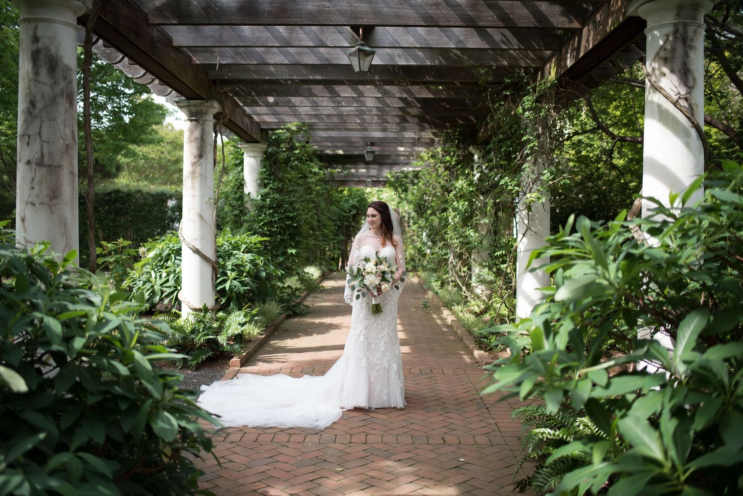 Elegant bride in white gown walks through a lush garden pergola holding her bouquet.
