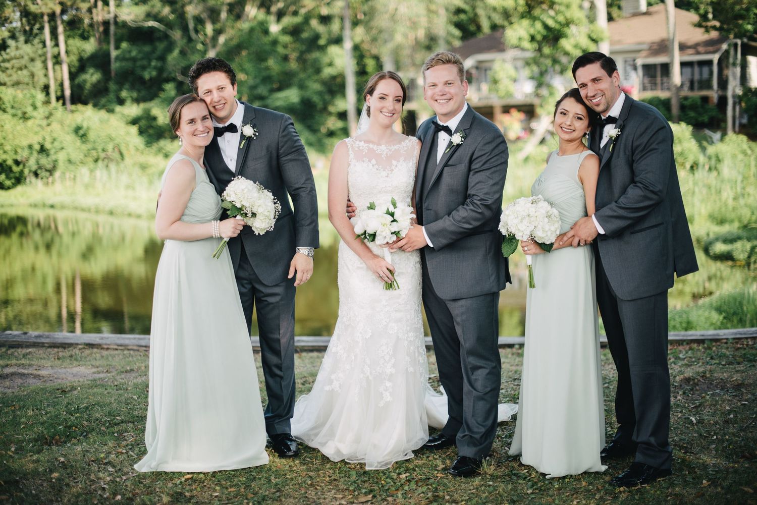 Wedding party of five poses elegantly in black tuxedos and sage bridesmaid gowns near a scenic pond.