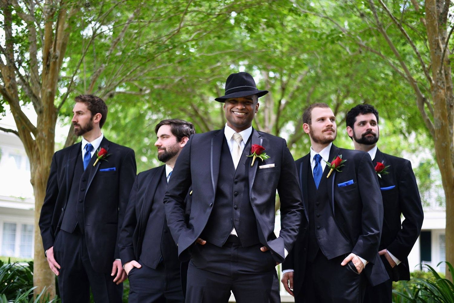 A groom in a black suit and fedora stands confidently with his groomsmen, all wearing dark suits with colorful boutonnieres.