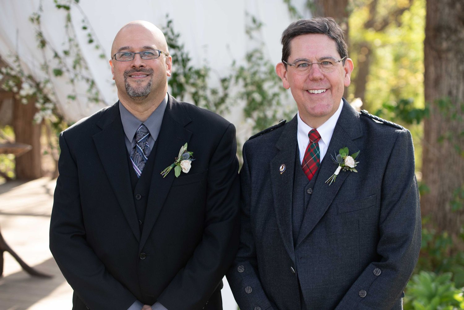 Two men in dark suits with boutonnieres smiling outdoors at a formal event or wedding ceremony.