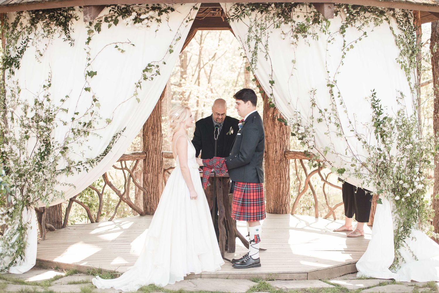 Bride and groom exchange vows under a rustic floral arch with white draping at an outdoor wedding ceremony.