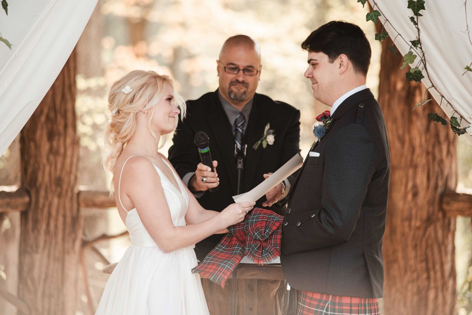 Outdoor wedding ceremony with bride, groom in kilt, and officiant under floral arch among trees.