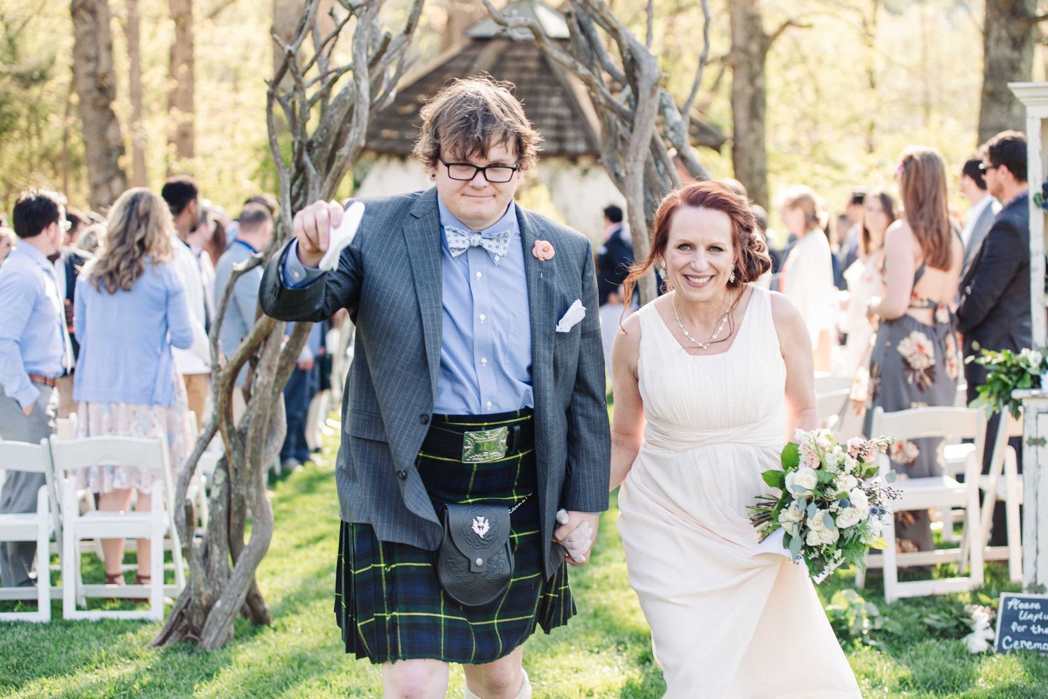 Newlywed couple exits outdoor wedding ceremony, groom in green tartan kilt, bride in white dress holding bouquet.