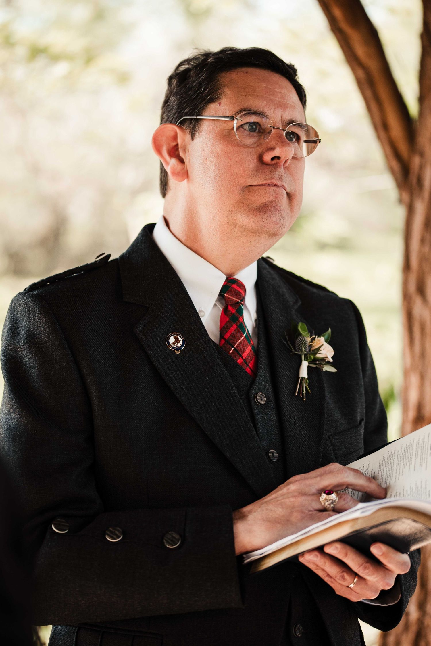 Man in dark suit with red plaid tie and boutonniere holds book outdoors, wearing glasses at formal event.