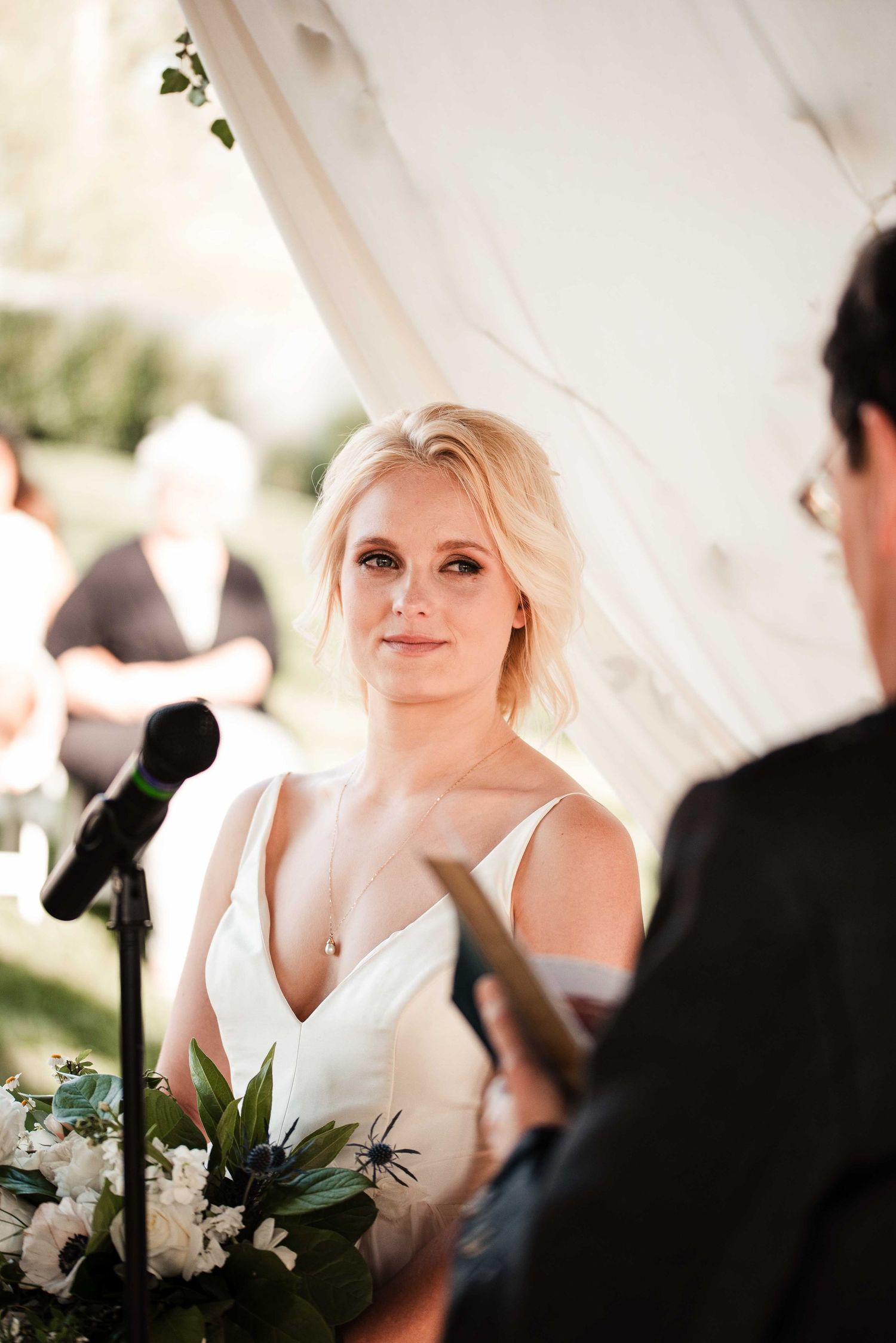 Blonde bride in white gown holds floral bouquet during outdoor wedding ceremony under a tent.
