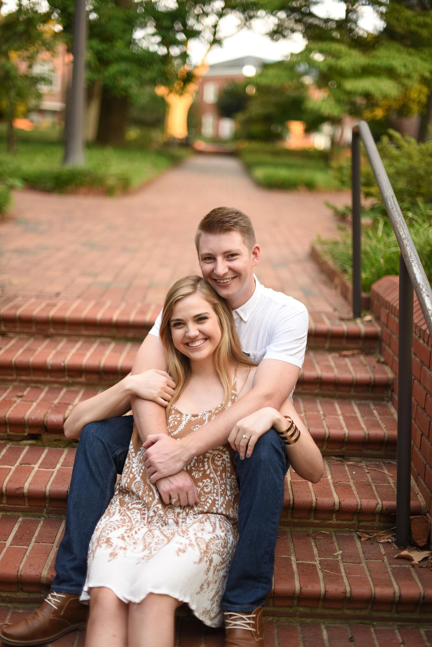 Happy couple posing on brick stairs outdoors, man hugging woman from behind with greenery behind.