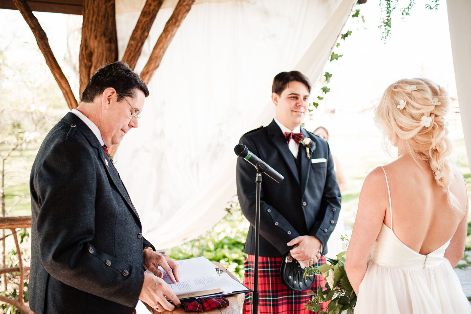 An officiant reads vows as a smiling groom in a tartan kilt stands with his bride at the altar.