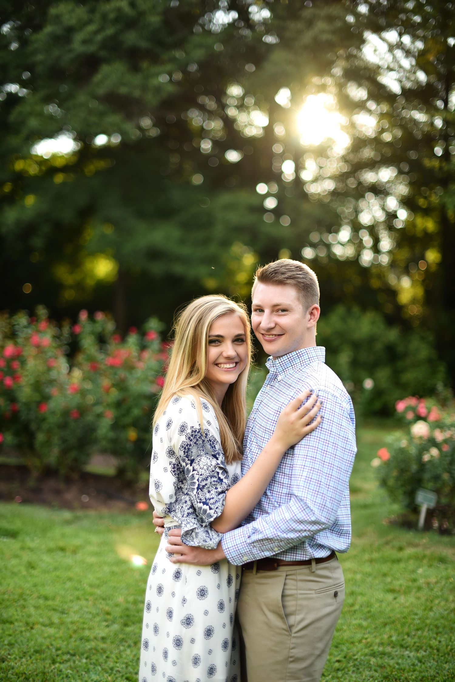 Couple embracing and smiling in a lush garden with blooming roses and golden sunlight.