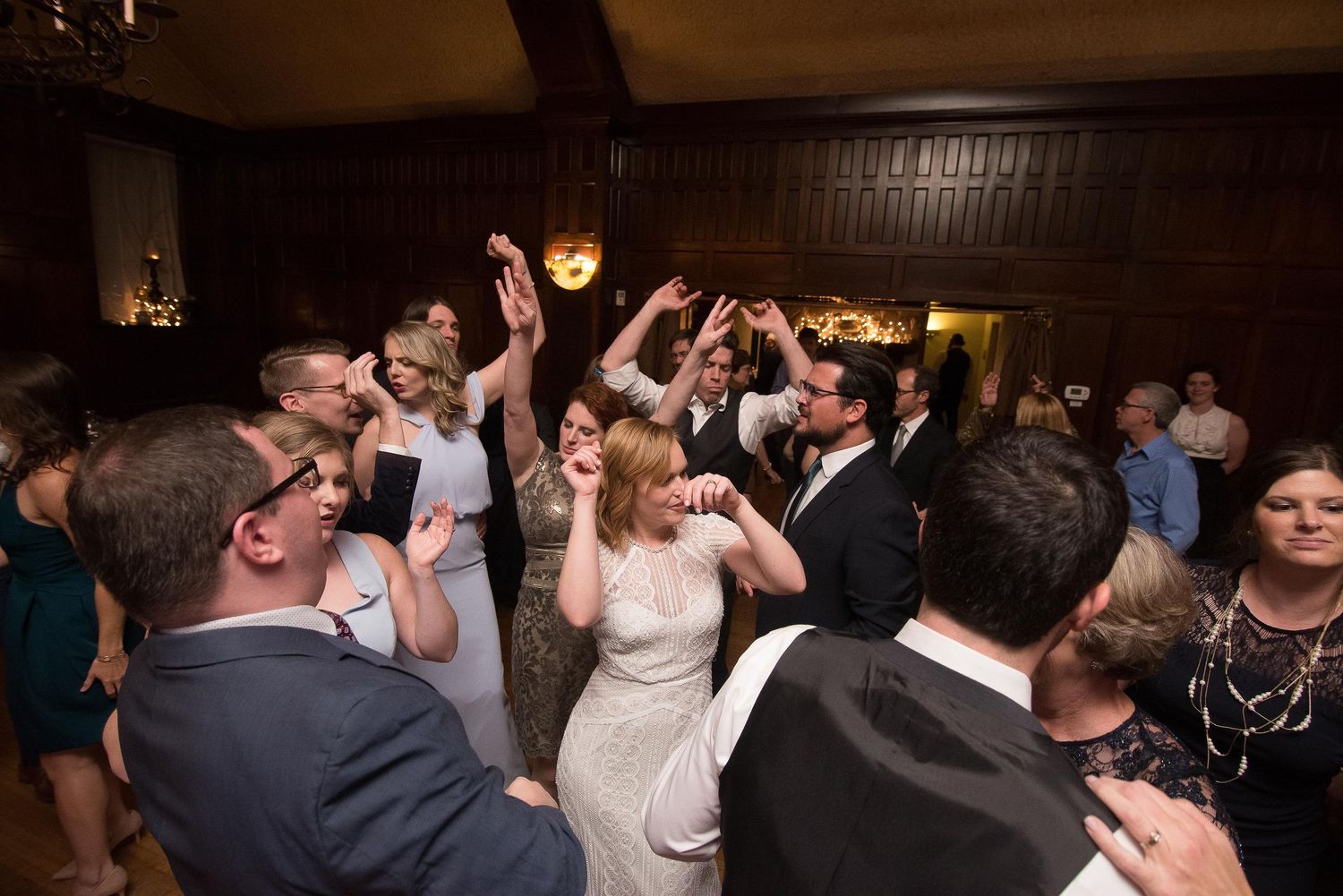 Wedding guests dancing joyfully at a lively indoor reception in a wood-paneled ballroom.