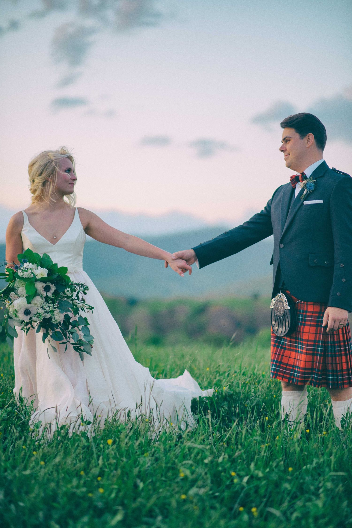 Bride in white gown holding hands with groom in red tartan kilt and navy jacket on a green field at sunset.