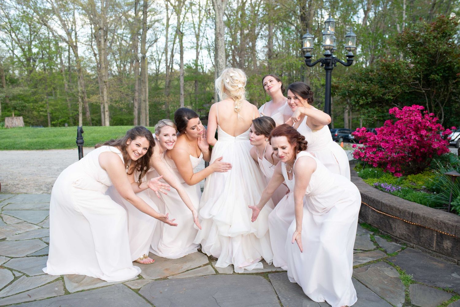 Bride and bridesmaids in white dresses posing outdoors on stone patio with blooming pink flowers and trees.