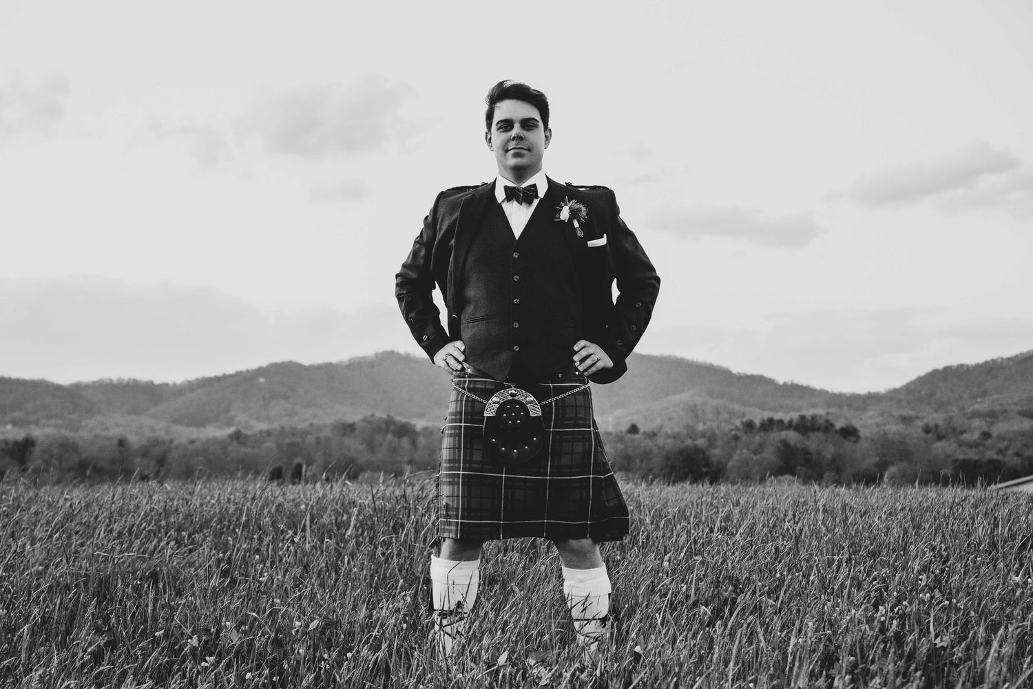 Man in traditional Scottish kilt and jacket standing confidently in a field with mountain backdrop, black and white photo.