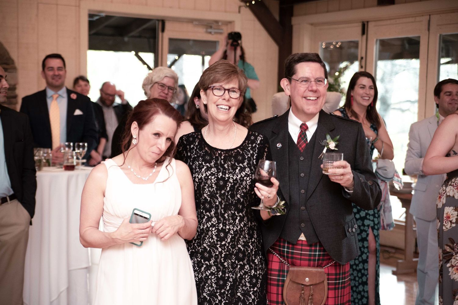 Wedding guests smiling and raising glasses during a toast, including a man in a red tartan kilt at a reception.