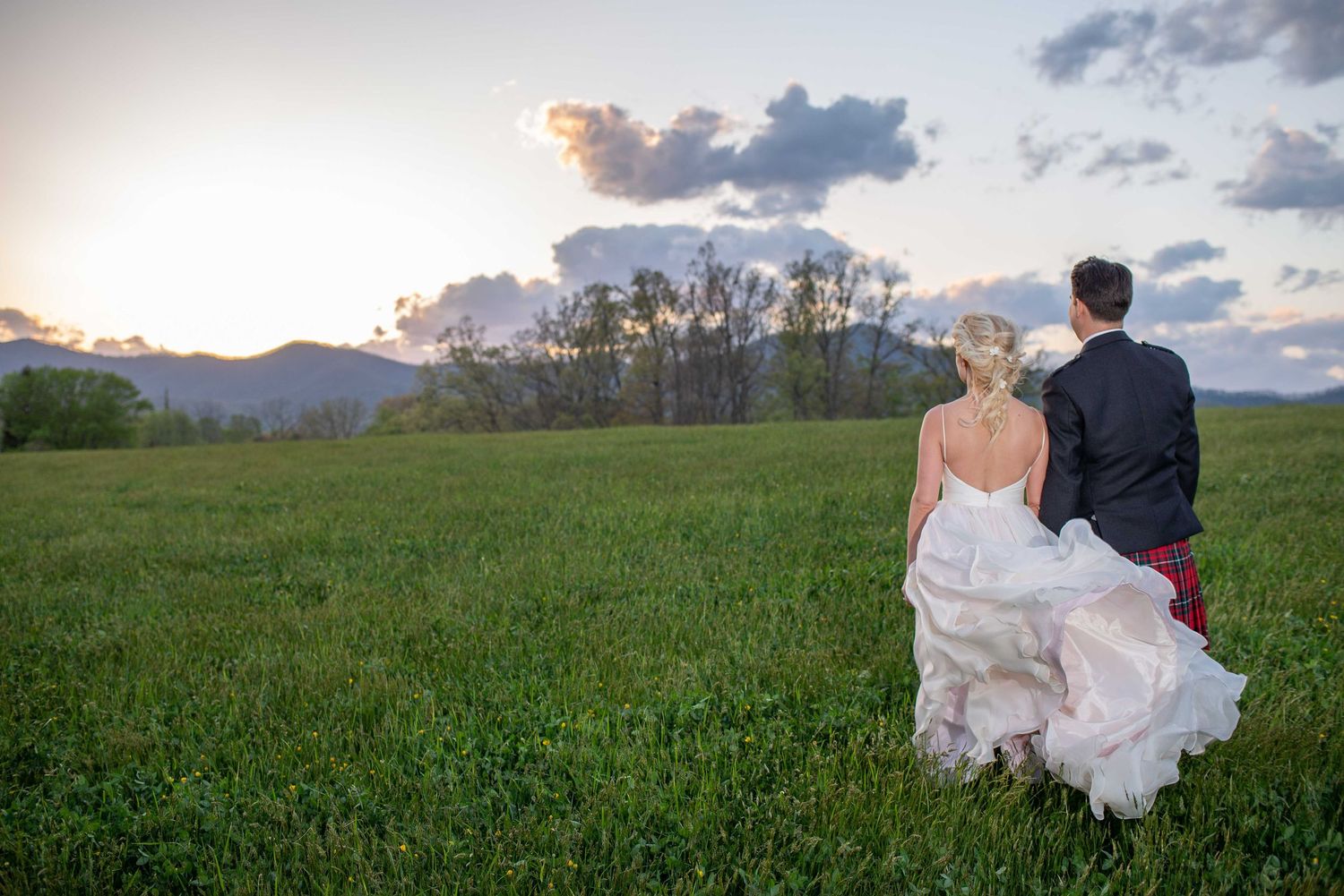 Bride and groom stand together in a lush green field at sunset with mountains in the background.