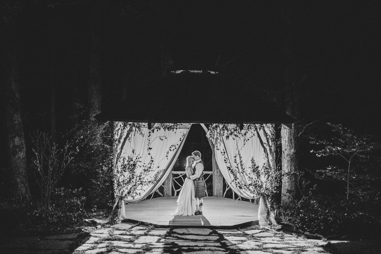 Black and white photo of a couple standing under a floral arch at an outdoor wedding ceremony.