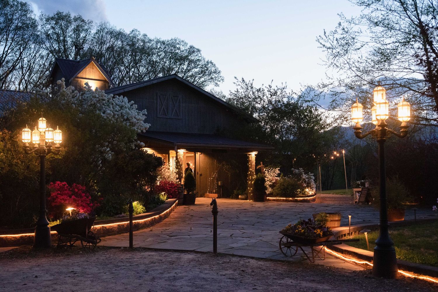 Elegant estate entrance illuminated at dusk with ornate lanterns, lush gardens, and snow-capped mountains in the background.