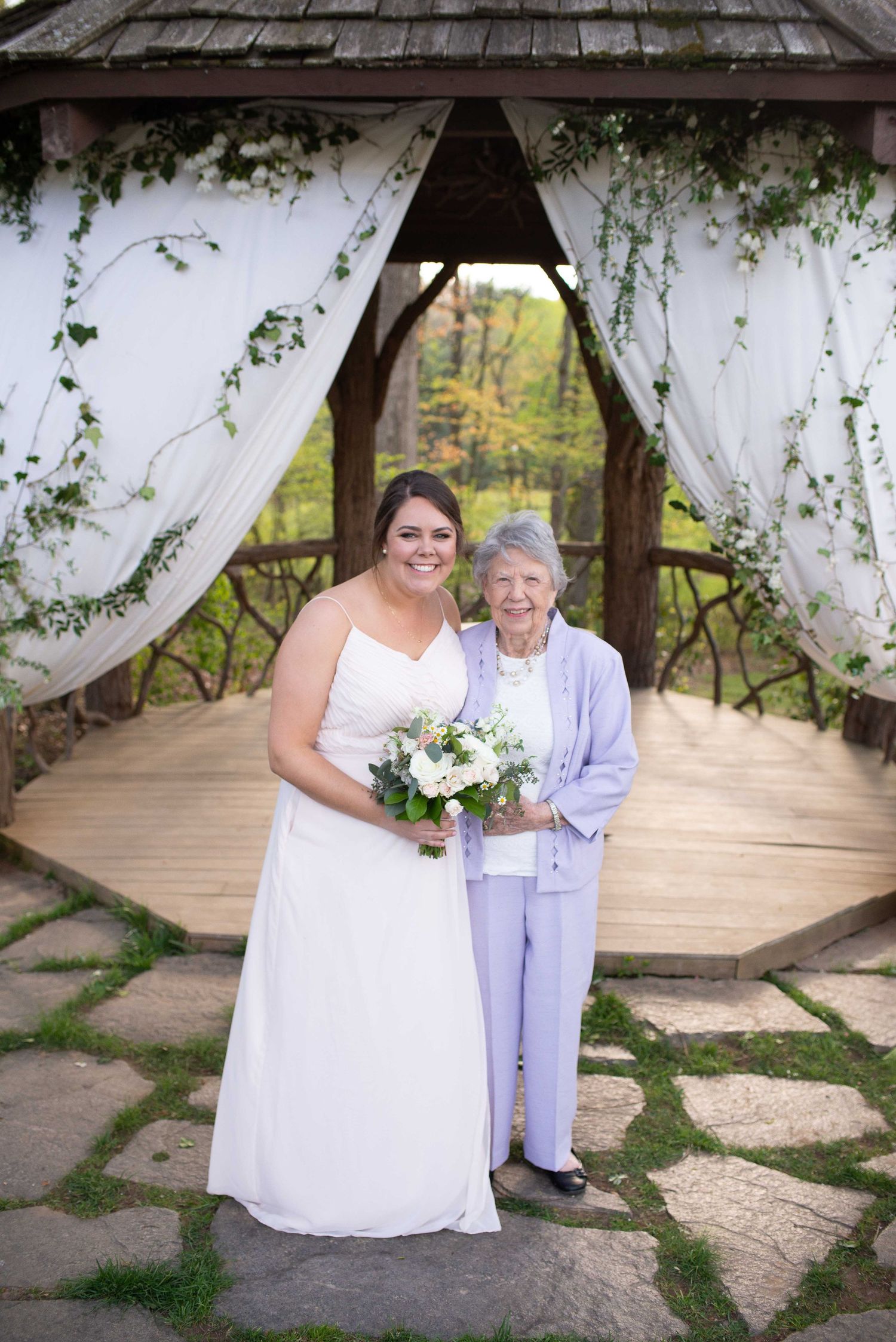 Bride in white gown holds bouquet beside elderly woman in lavender suit under decorated gazebo at outdoor wedding.