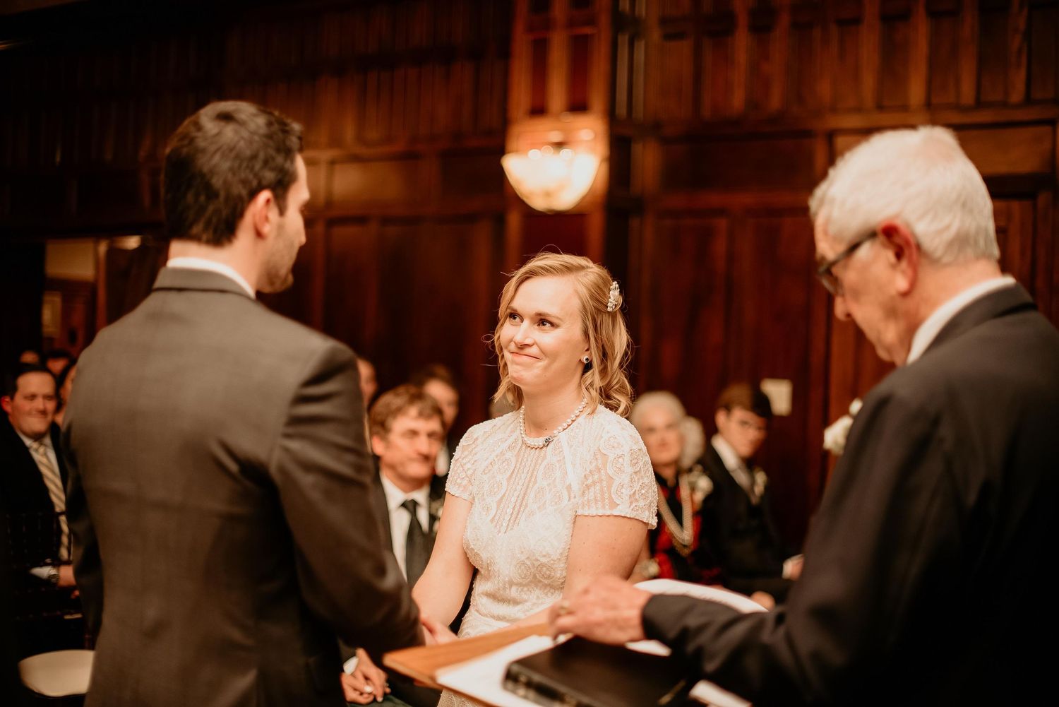 Bride smiling joyfully at groom during indoor wedding ceremony with guests and officiant in elegant wood-paneled venue.