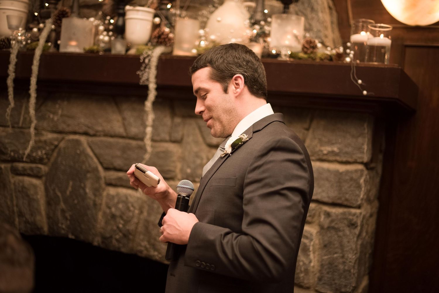 Man in gray suit reading a card by a stone fireplace decorated with holiday greenery and candles.