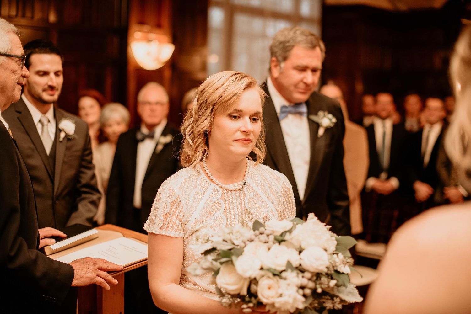 Bridesmaid holding white floral bouquet during wedding ceremony in elegant wood-paneled venue.