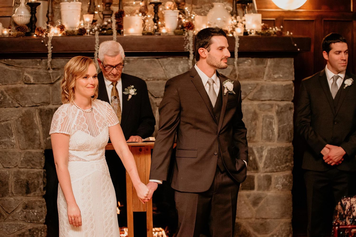 Bride and groom hold hands at rustic stone fireplace wedding ceremony with officiant standing behind them.