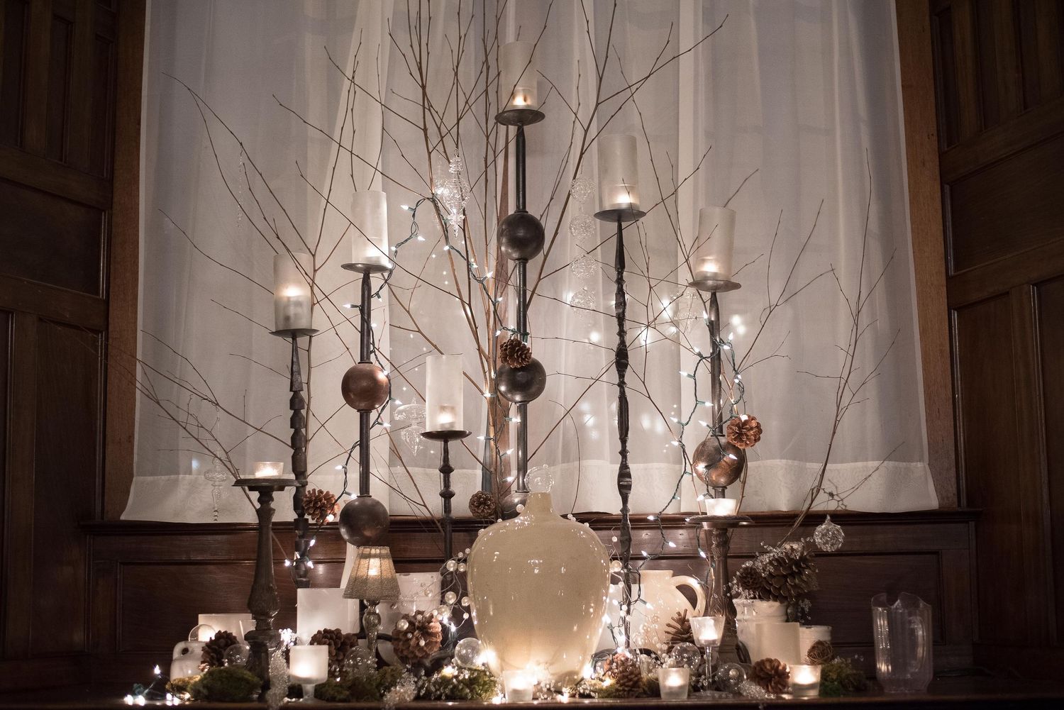 Elegant winter centerpiece with tall candlesticks, white birch branches, ornaments, and glowing candles on a wooden sideboard.