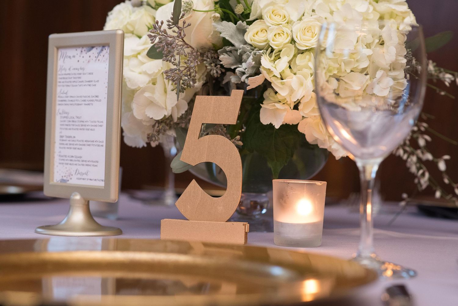 Elegant wedding table setting with gold number 5, white floral centerpiece, candle, and glassware.