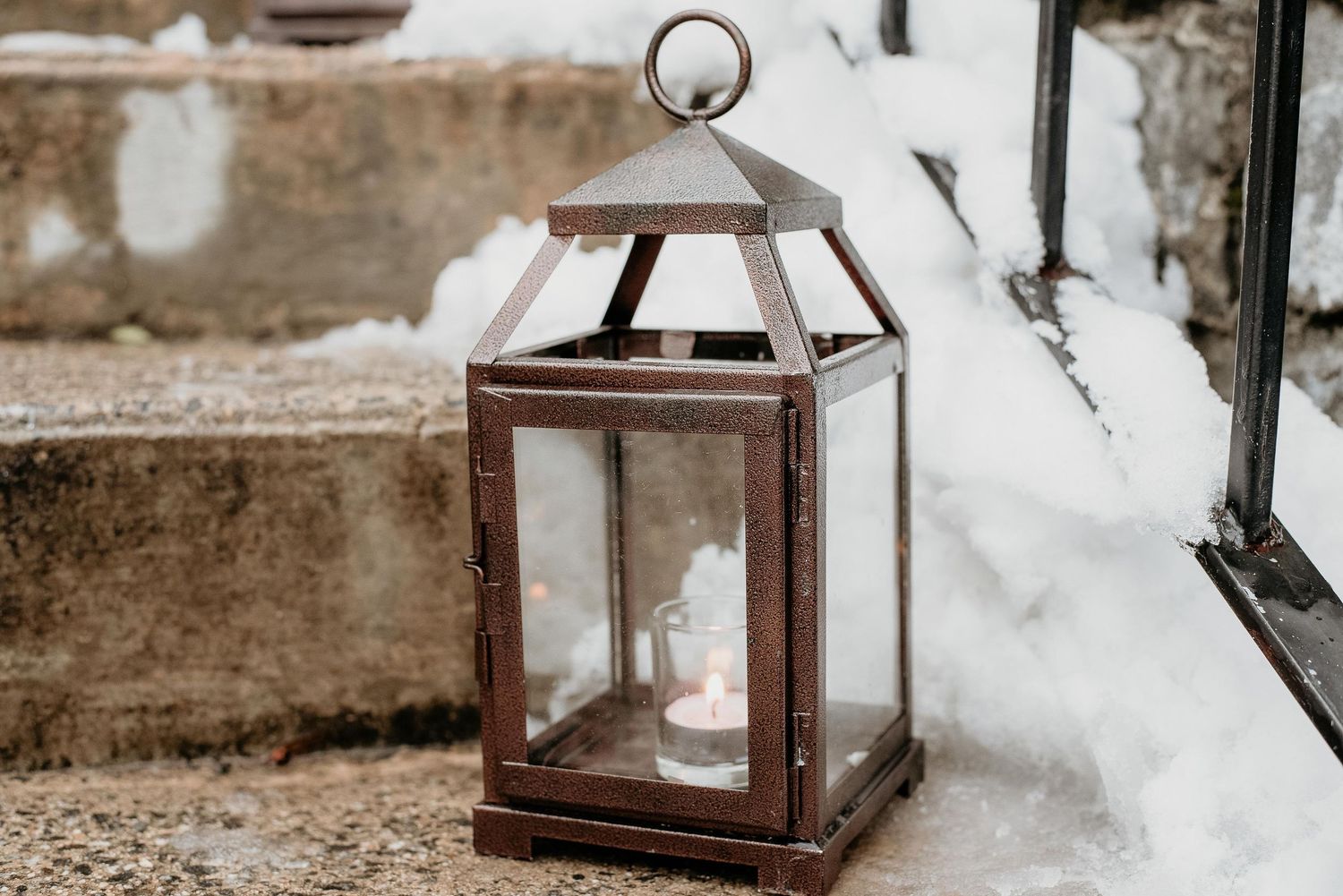 Rustic metal lantern with glowing candle sitting on stone steps surrounded by snow in winter.