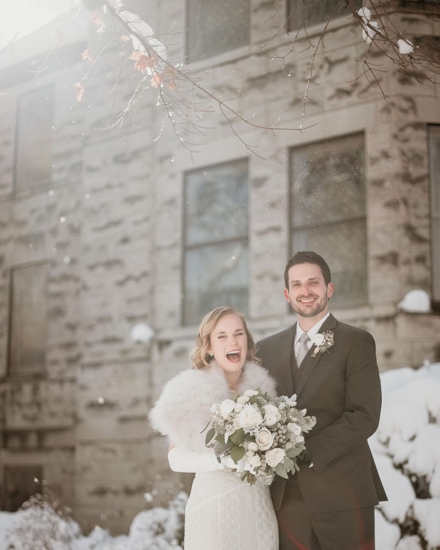 Happy couple poses in snow outside stone building, bride in white fur wrap holding bouquet, groom in gray suit.