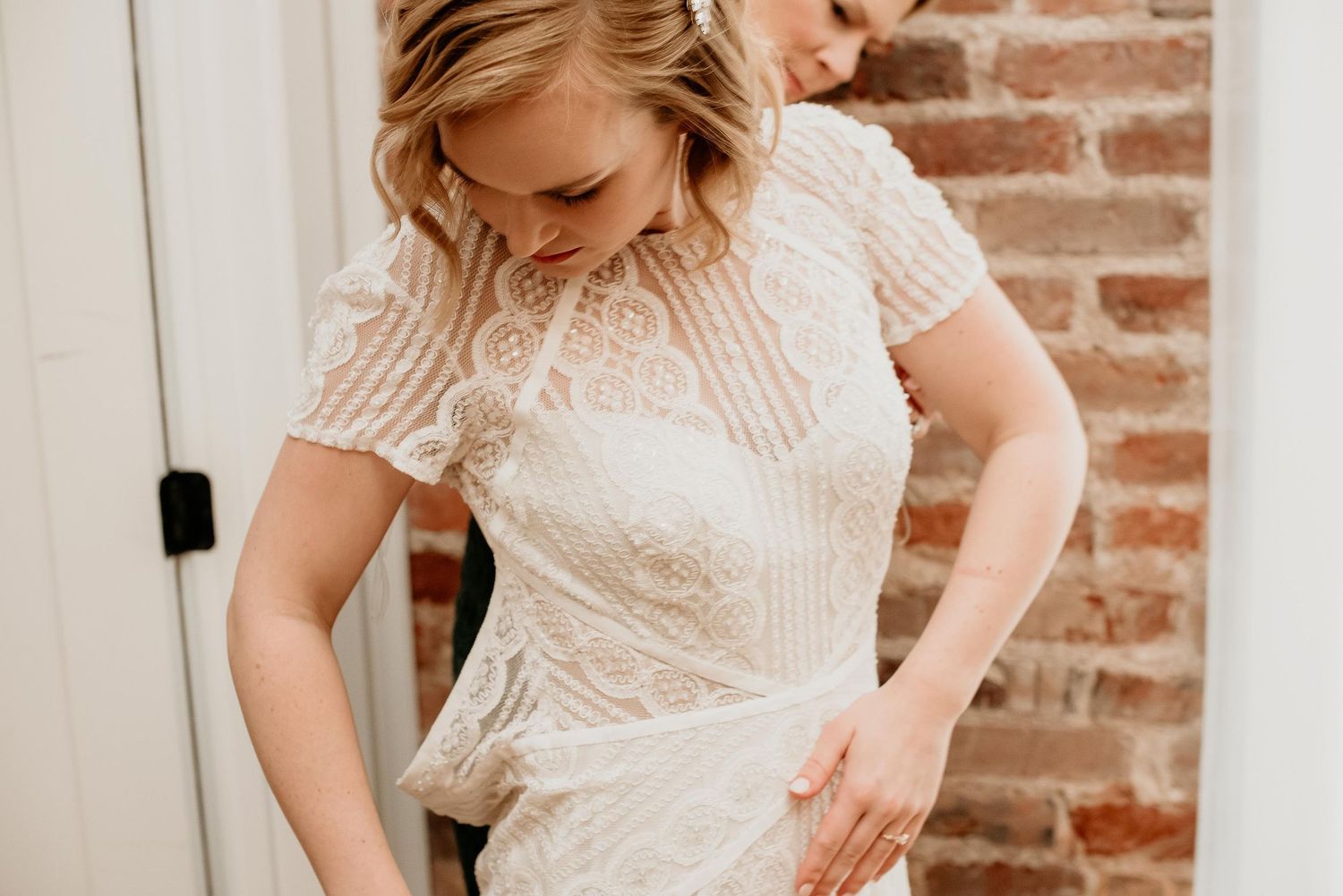 Bride in elegant lace wedding gown with short sleeves being helped with dress near rustic brick wall.