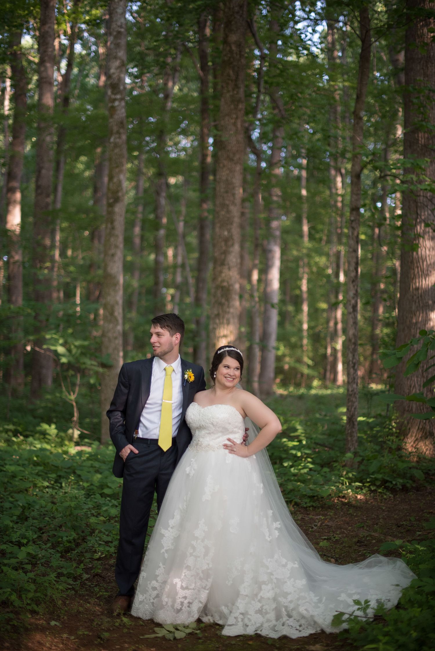 Bride and groom posing in lush forest, groom in navy suit with yellow tie, bride in lace gown.