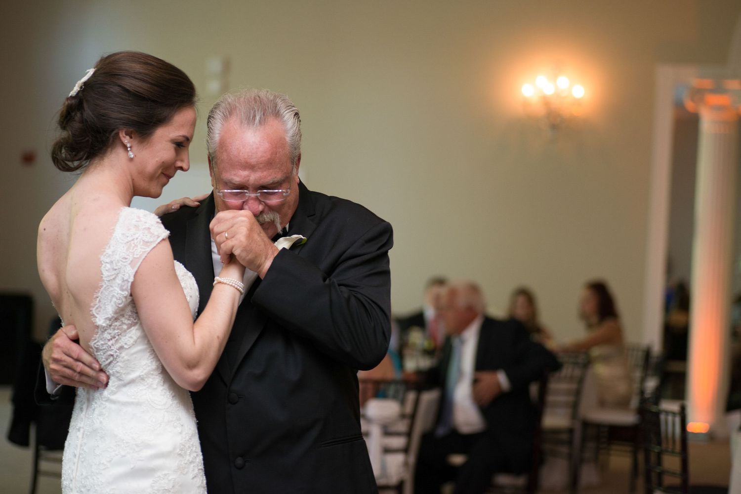 Emotional father wipes tears while sharing a touching father-daughter dance with bride at wedding reception.