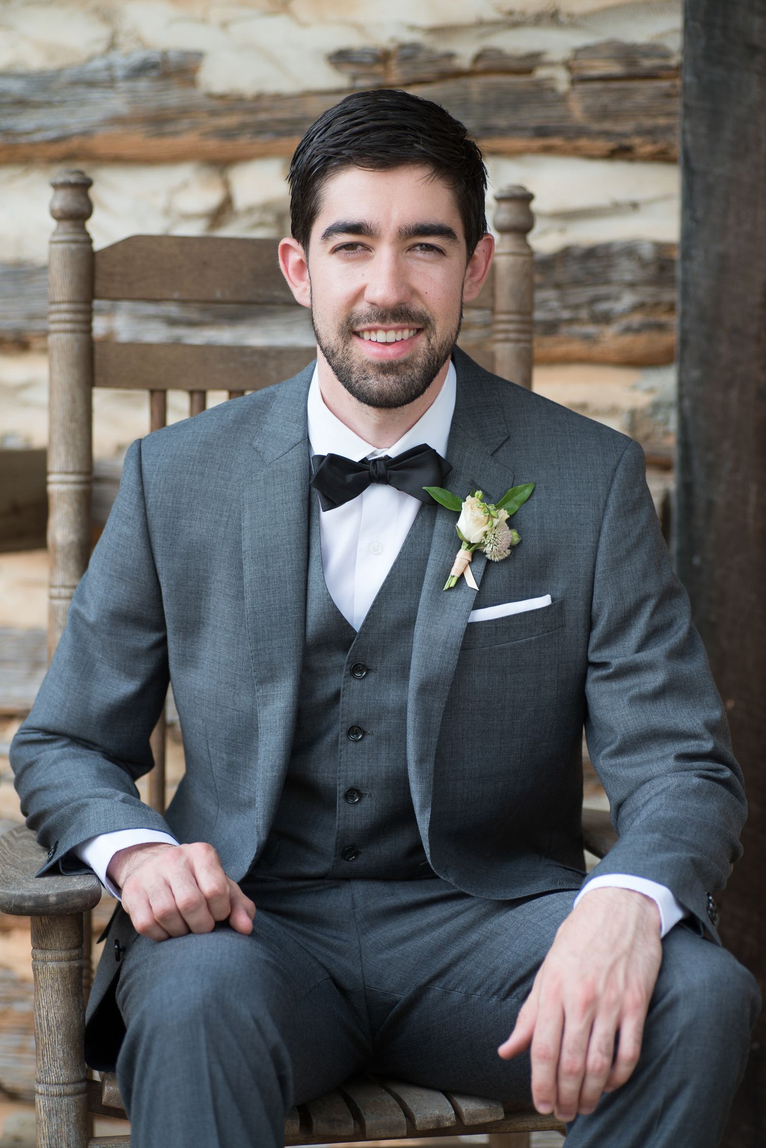 Groom in gray suit with bow tie and boutonniere seated outdoors.