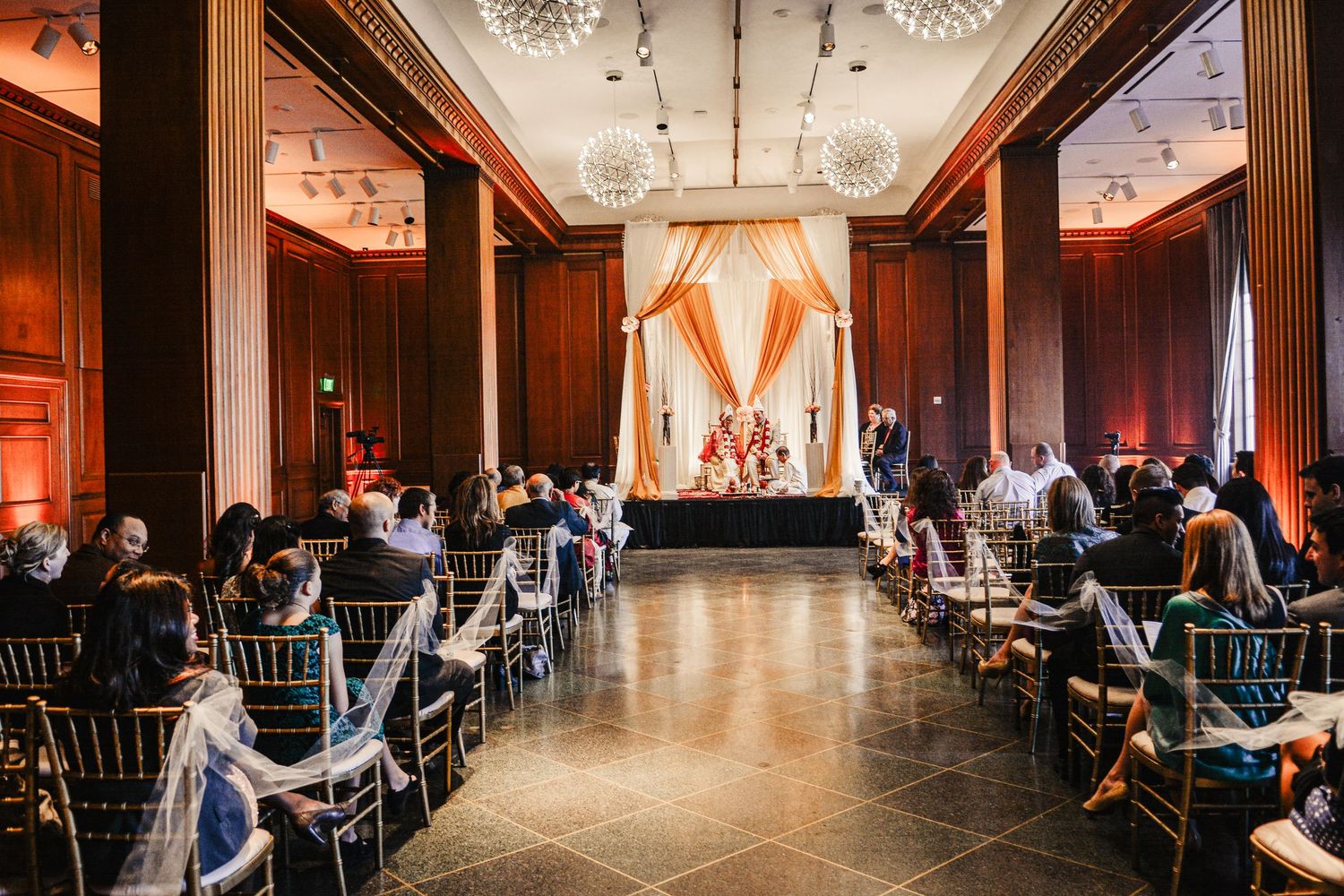 Elegant indoor wedding ceremony with golden draping, chandelier lighting, and guests seated in a grand hall.