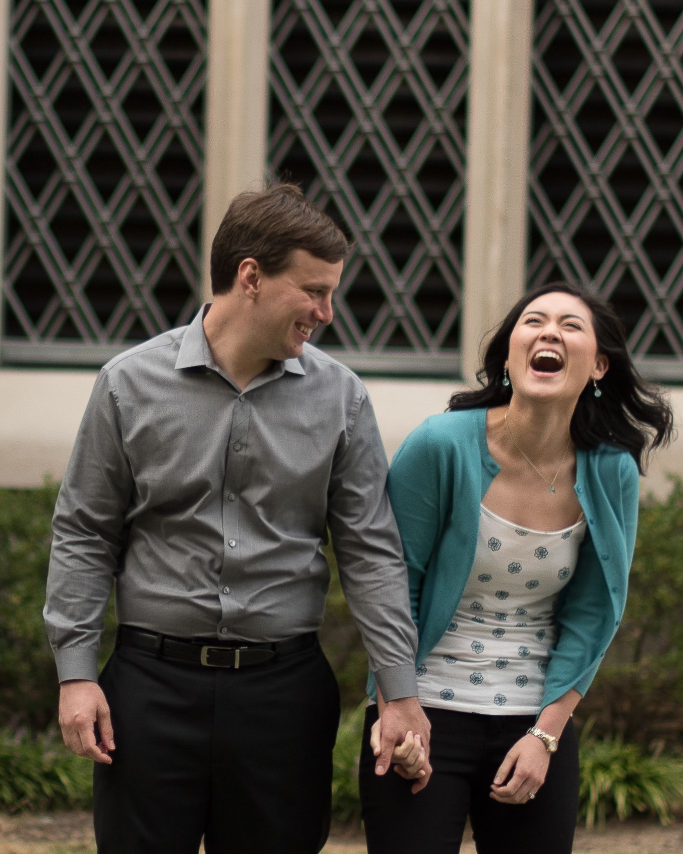 Joyful couple laughing and holding hands outdoors near decorative lattice window architecture.