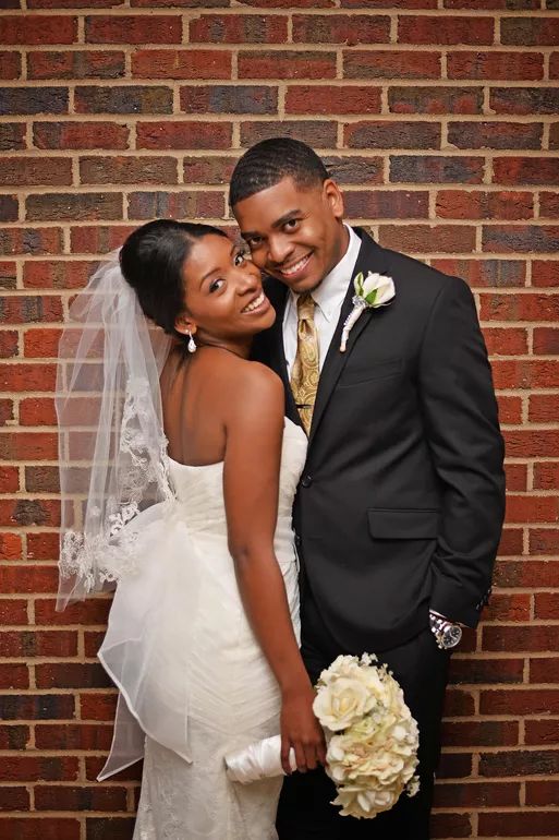 Smiling African American bride and groom pose together against a red brick wall.