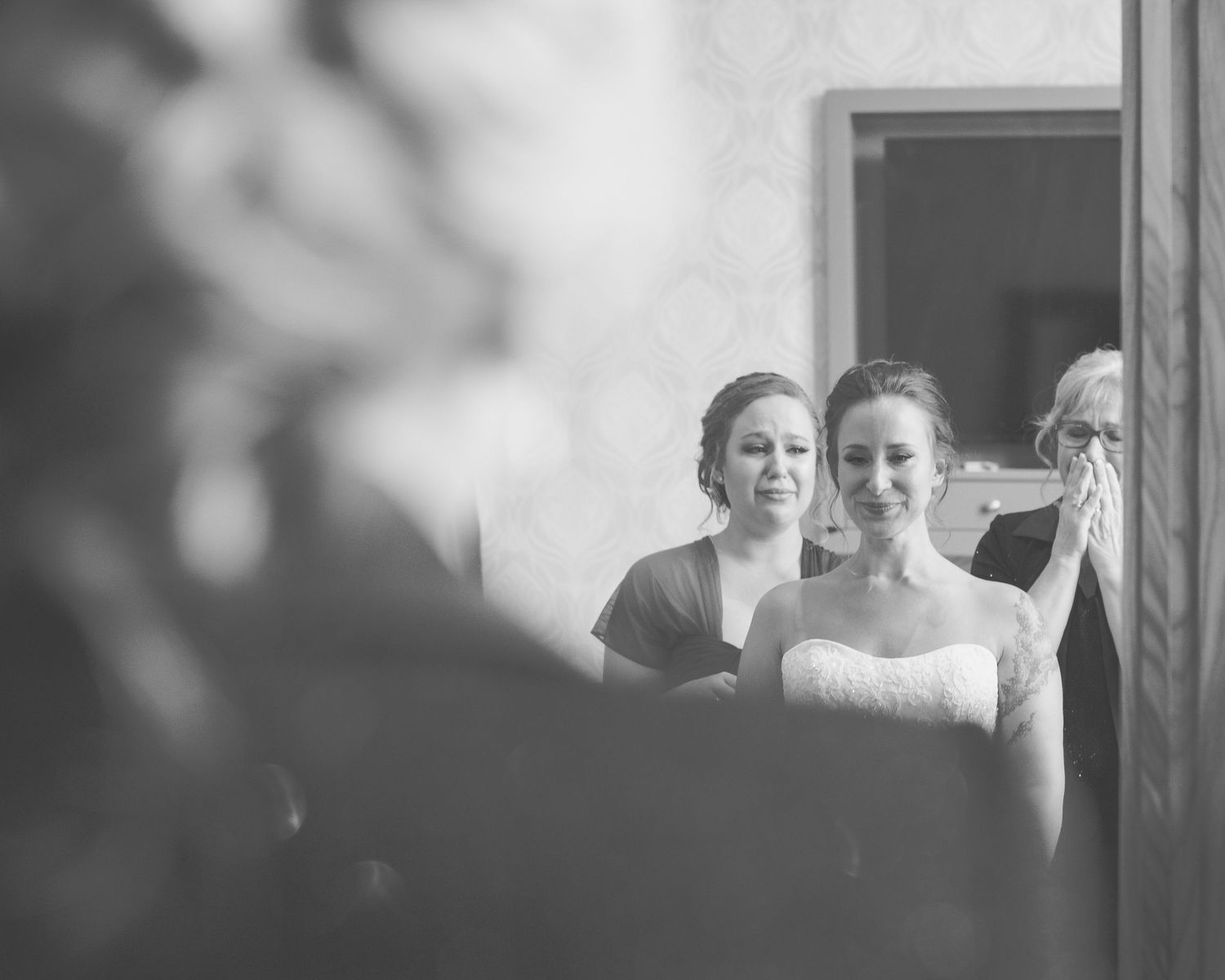 Emotional black and white photo of bride smiling with bridesmaid and tearful family member.