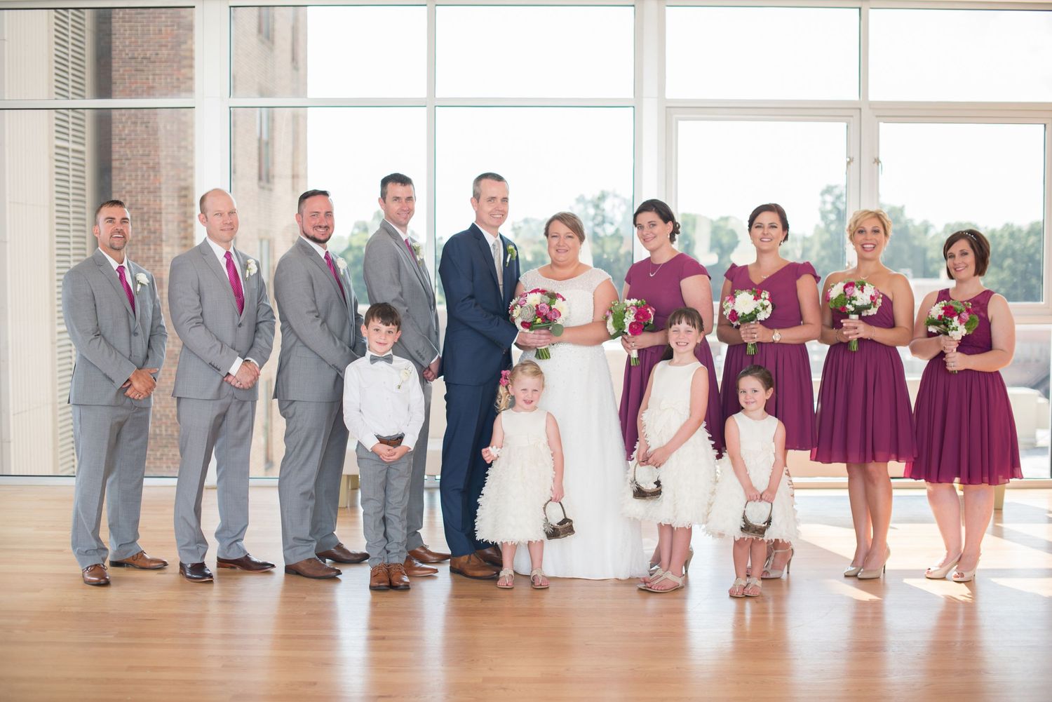 Wedding party posing indoors with bridesmaids in purple dresses and groomsmen in gray suits.