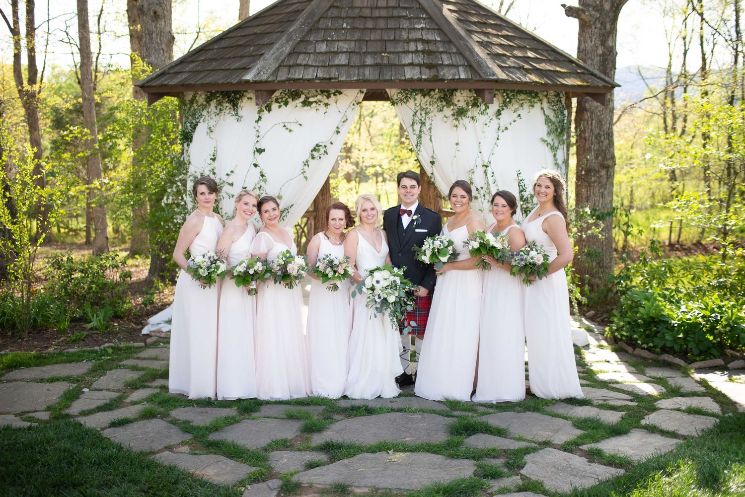 A wedding party poses under a floral gazebo with bridesmaids in blush gowns holding white bouquets.