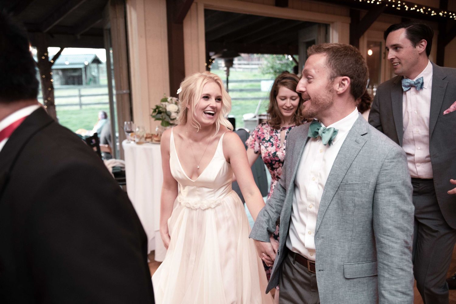 Smiling bride in a white gown holds hands with groom in gray suit and teal bow tie at their wedding reception.