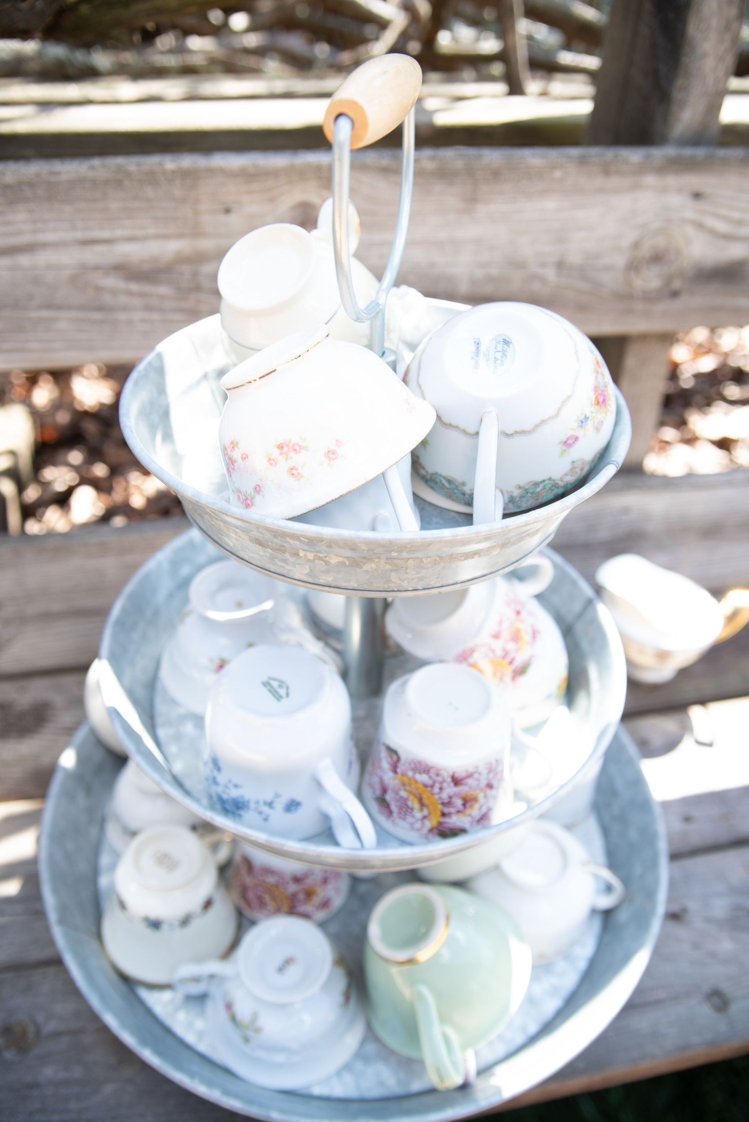 Three-tier galvanized metal stand displaying vintage floral teacups and saucers at an outdoor rustic wedding reception.