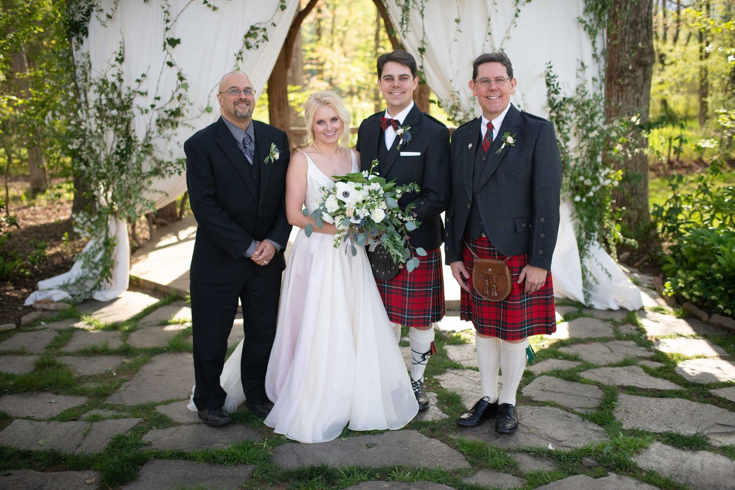 Bride and groom in tartan kilt pose with two men in formal attire before white floral wedding arch.