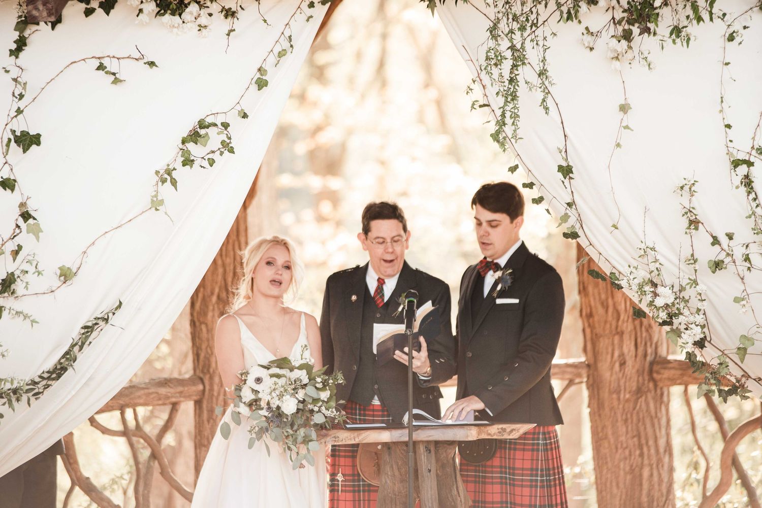 Outdoor wedding ceremony with bride, groom in tartan kilt, and officiant under floral arch with white draping.