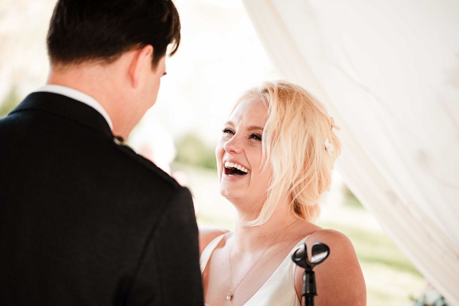 Bride laughing joyfully at groom during outdoor wedding ceremony under white tent.