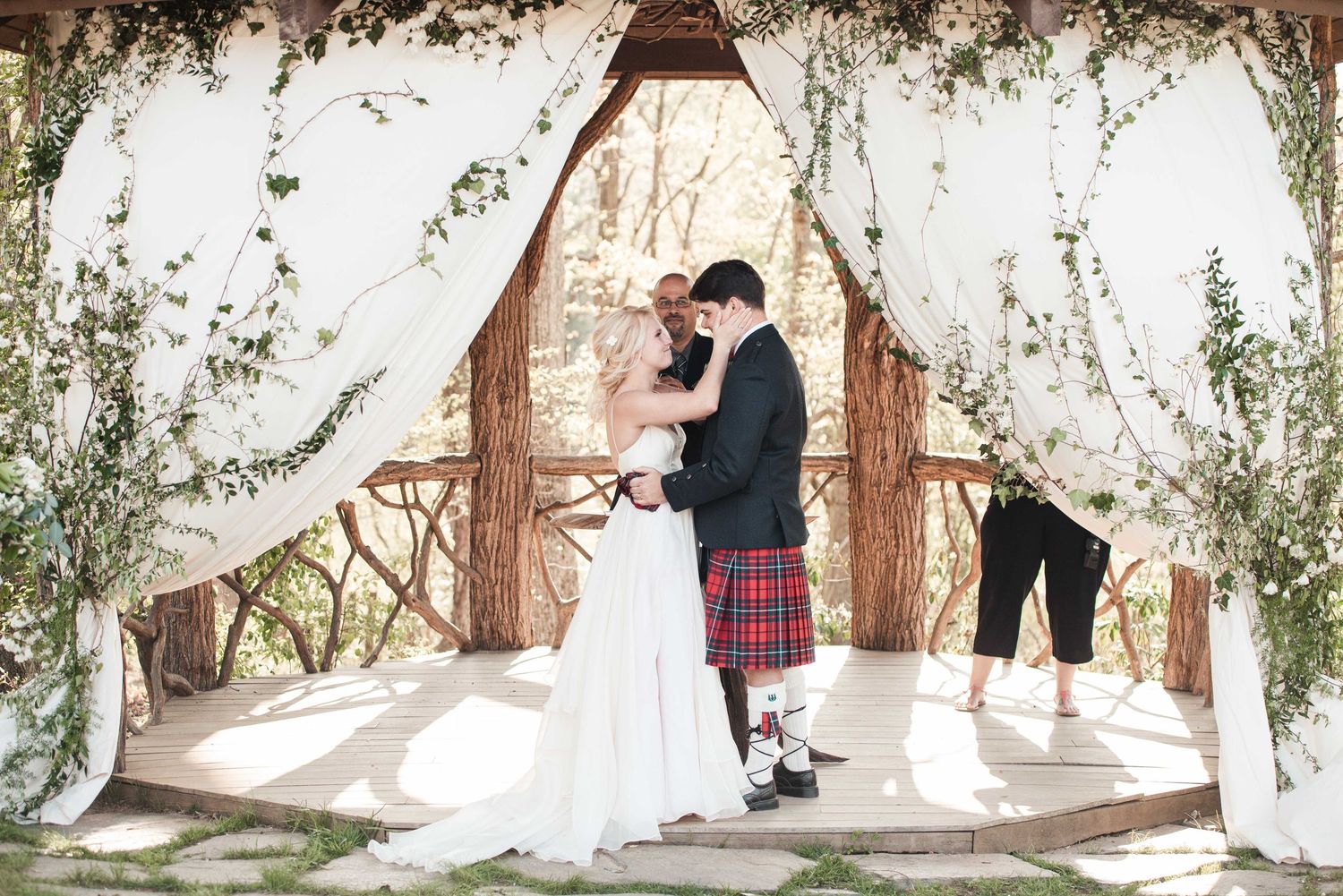 Bride and groom share first kiss under floral arch with white draping at outdoor wedding ceremony.