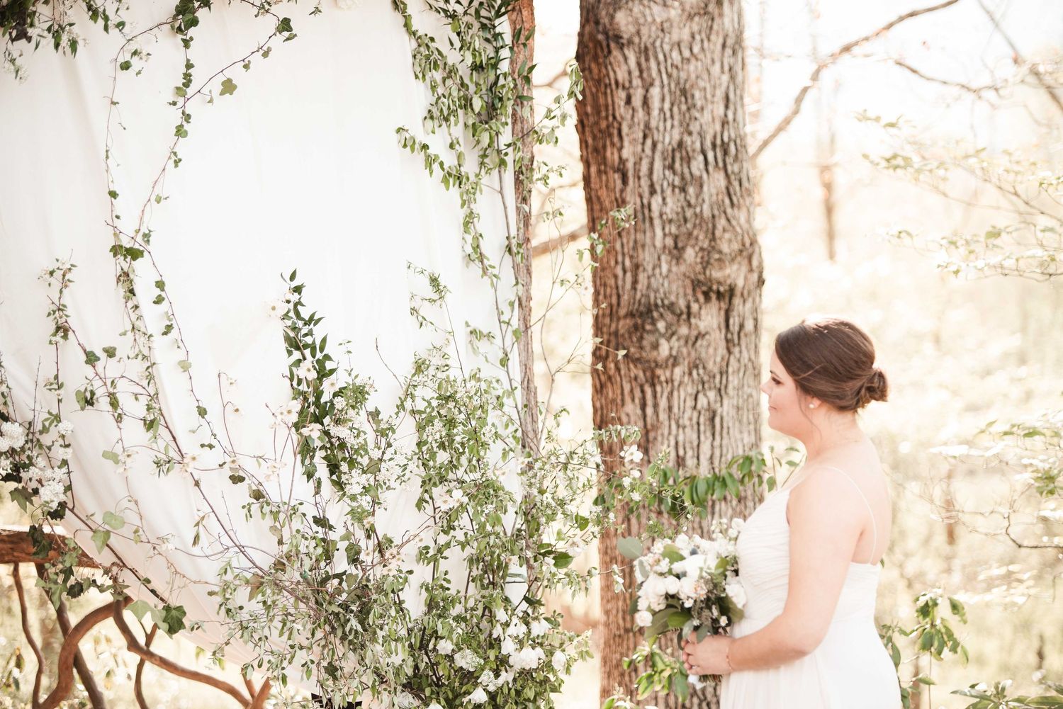 Bride in white gown holding bouquet stands beside a floral arch with greenery and white flowers in an outdoor woodland setting.